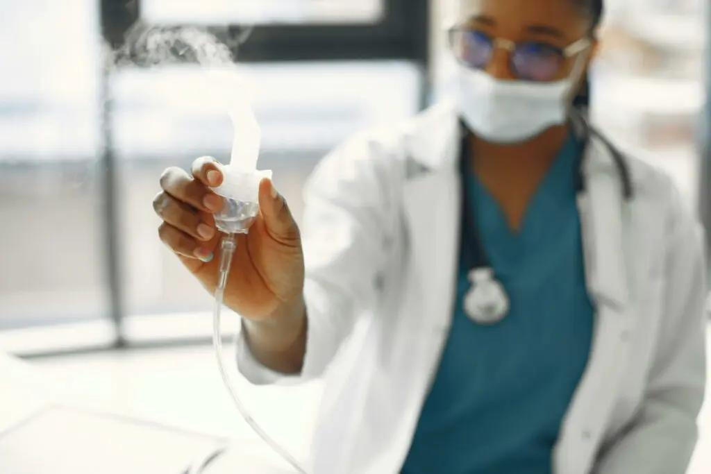 A healthcare professional in scrubs and a lab coat holds a nebulizer used for respiratory therapy, demonstrating proper use of respiratory care equipment in a clinical setting.